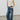 A model wearing a beige ruched sleeveless top and wide-leg blue denim jeans, standing against a plain light gray wall and concrete floor.