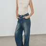 A model wearing a beige ruched sleeveless top and wide-leg blue denim jeans, standing against a plain light gray wall and concrete floor.