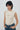 a beige ruched sleeveless top and blue jeans, standing against a light gray studio background.