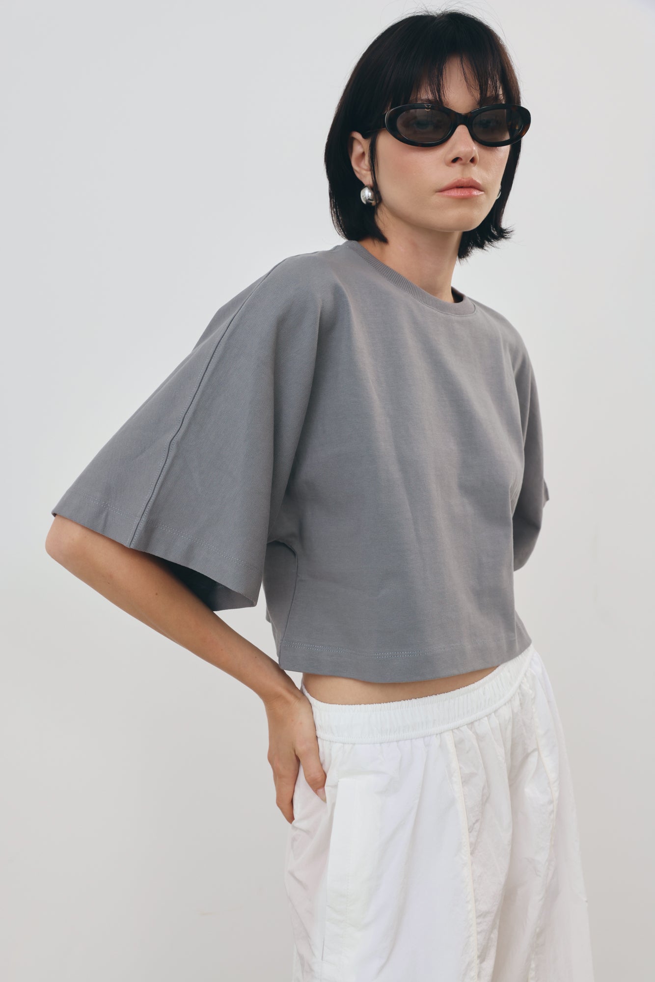 a gray cropped t-shirt with wide, voluminous sleeves and white pants, styled with black sunglasses against a plain light gray background.