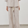 A person wearing a light beige matching two-piece outfit with a short-sleeve button-front top and wide-leg pants, standing against a plain light gray wall and floor.