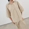 A model wearing an oversized beige short-sleeve button-up shirt and matching wide-leg trousers, styled with dark sunglasses and black shoes, standing in a minimalist room with a white wall and gray floor.