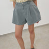 A person wearing high-waisted, wide-leg denim shorts in a faded light blue wash, paired with a cream shirt and dark loafers, standing against a plain light gray wall and floor.