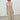 A model wearing a saga smocked midi dress with thin spaghetti straps, styled with black sunglasses, gold hoop earrings, and black leather loafers, standing against a minimalist white wall and gray floor.