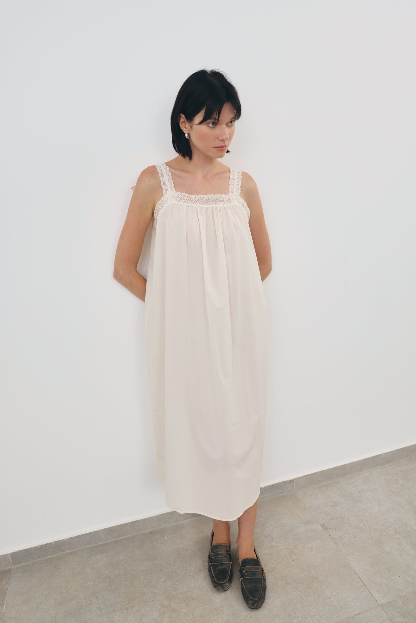 A woman with short black hair stands against a white wall wearing a cream-colored linen slip dress with lace trim on the straps and square neckline, paired with dark loafers.
