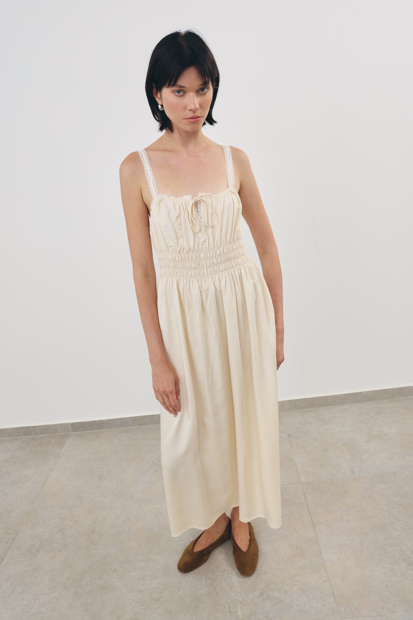 A model wearing a cream-colored viscosa midi dress with a smocked bodice and thin lace shoulder straps, paired with khaki flat shoes, standing in a minimalist studio setting.