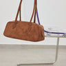 A brown suede duffle bag with dual top handles, hanging from a clear acrylic chair with a purple cushion on a metal cantilever chair in a minimalist room.